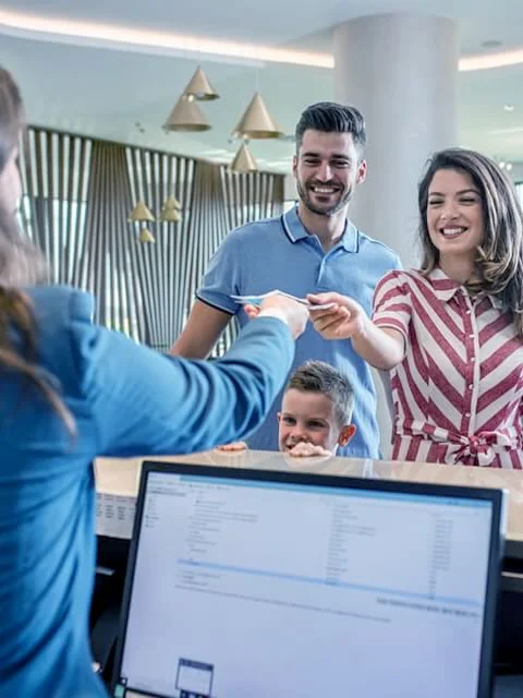 A imagem mostra uma recepção de uma pousada organizada e uma família feliz fazendo checkin